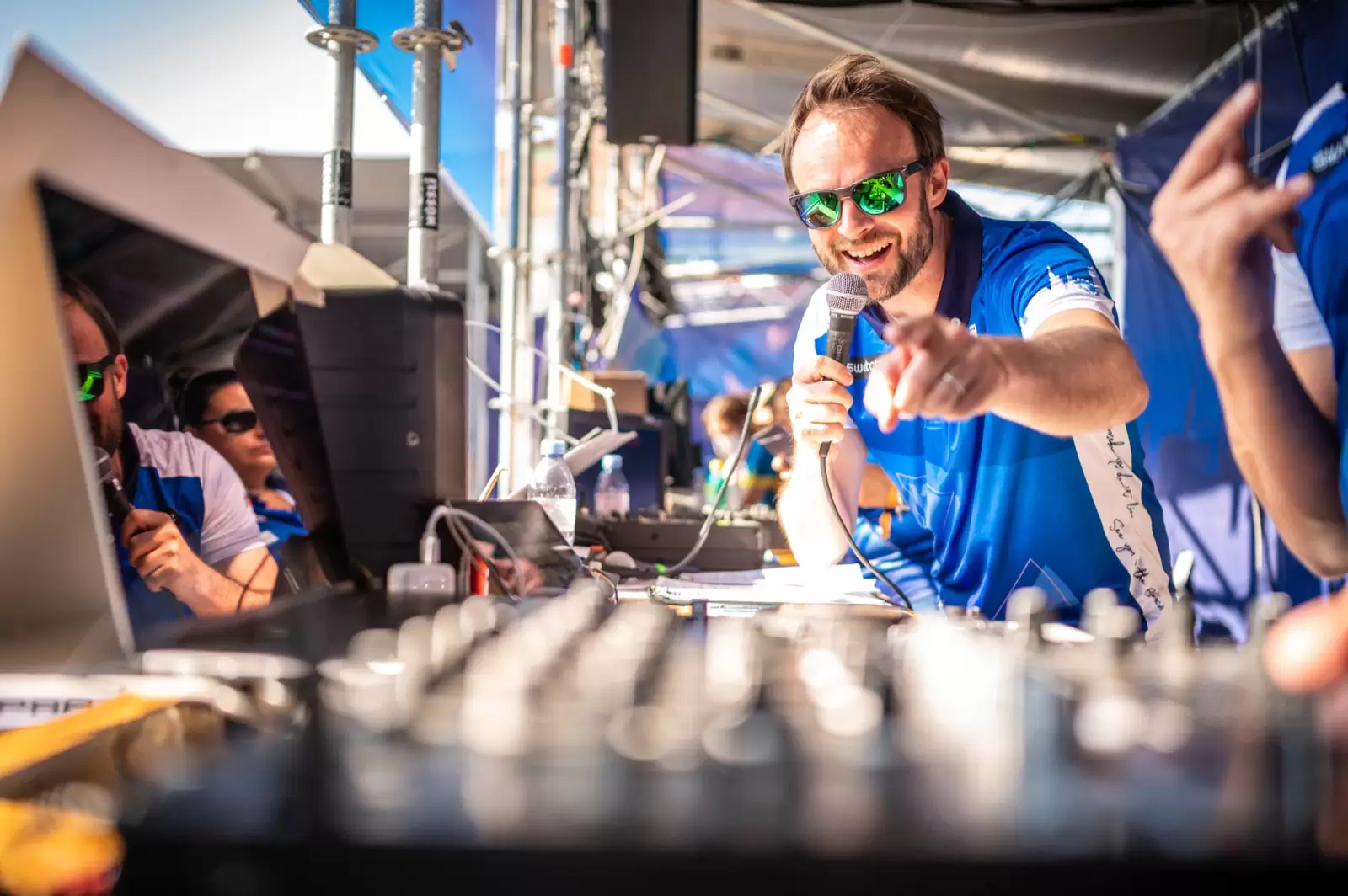 Florian Rudig mit Sonnenbrille und Mikrofon am Mischpult der Stadionregie, er zeigt lachend auf das Publikum