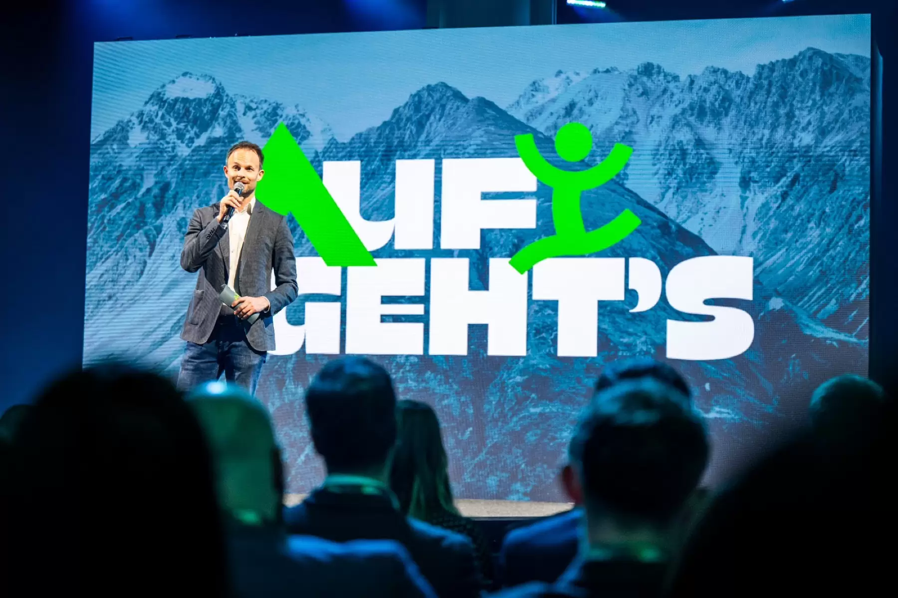 Florian Rudig moderiert die Merkur Jahresgala 2025 vor einer riesigen Leinwand mit Bergpanorama und dem Schriftzug Auf geht’s