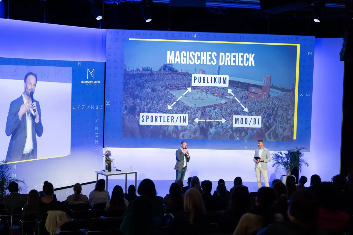 Keynote von Florian Rudig auf großer Bühne, auf der Leinwand das magische Dreieck aus Publikum, Sportlern und Moderation Keynote von Florian Rudig auf großer Bühne, auf der Leinwand das magische Dreieck aus Publikum, Sportlern und Moderation