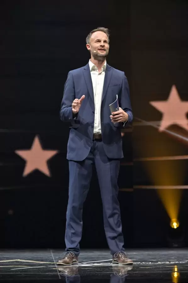 Florian Rudig im blauen Anzug mit Moderationskarte auf der Bühne beim OVB Europatag 2025, hinter ihm leuchtende Sterne Florian Rudig im blauen Anzug mit Moderationskarte auf der Bühne beim OVB Europatag 2025, hinter ihm leuchtende Sterne